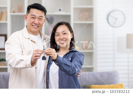 Happy young Asian family, man and woman standing hugging in house, apartment and holding keys in hands, pointing at camera, smiling. Happy young Asian family, man and woman standing hugging in house, apartment and holding keys in hands, pointing at camera, smiling. 101583212