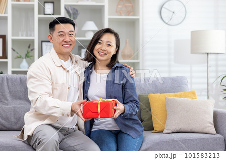 A young Asian couple, a man and a woman, are sitting on the sofa at home and holding a gift box in their hands. Smiling and happy looking at the camera. A young Asian couple, a man and a woman, are sitting on the sofa at home and holding a gift box in their hands. Smiling and happy looking at the camera. 101583213