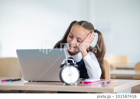 Caucasian girl sits at a desk at school and studies at a laptop. Alarm clock on the table. Caucasian girl sits at a desk at school and studies at a laptop. Alarm clock on the table. 101587416