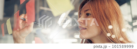 Businesswoman writing on sticky notes on glass wall while working in office 101589999