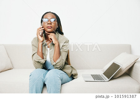 African American woman business freelancer working sitting on the couch at home in a laptop and phone, business calls and correspondence sadness and anger, home clothes and eyeglasses, light interior African American woman business freelancer working sitting on the couch at home in a laptop and phone, business calls and correspondence sadness and anger, home clothes and eyeglasses, light interior 101591387