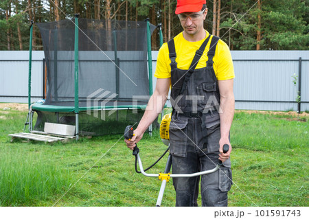 A male gardener mows the green grass of the lawn in the backyard with a gasoline mower. Trimmer for the care of a garden plot 101591743