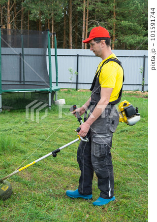 A male gardener mows the green grass of the lawn in the backyard with a gasoline mower. Trimmer for the care of a garden plot A male gardener mows the green grass of the lawn in the backyard with a gasoline mower. Trimmer for the care of a garden plot 101591744