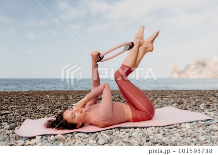 Woman sea pilates. Sporty happy middle aged woman practicing fitness on beach near sea, smiling active female training with ring on yoga mat outside, enjoying healthy lifestyle, harmony and meditation 101593738