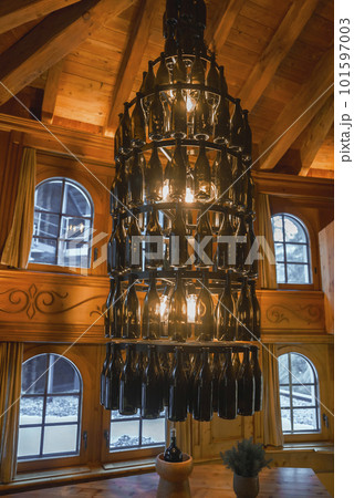 Illuminated chandelier made of empty alcohol bottles hanging on wooden ceiling in alpine chalet with windows in background, luxury travel concept for tourists Illuminated chandelier made of empty alcohol bottles hanging on wooden ceiling in alpine chalet with windows in background, luxury travel concept for tourists 101597003