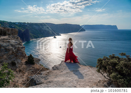 A girl with flowing hair in a long red dress stands on a rock above the sea. The stone can be seen in the sea. Sunny path to the sea from the sun. A girl with flowing hair in a long red dress stands on a rock above the sea. The stone can be seen in the sea. Sunny path to the sea from the sun. 101600781