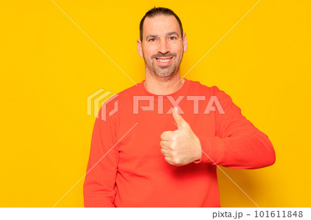 Bearded Hispanic man in his 40s wearing a red jumper proudly raising his thumb up while looking and smiling at the camera, isolated on yellow studio background. 101611848