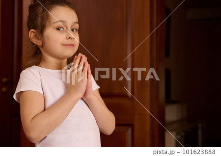 Close-up portrait of a 5-6 years old Caucasian little pretty child girl in white t-shirt, keeping hands palms together while practicing yoga, smiling looking at camera. Cute kid meditating indoors 101623888