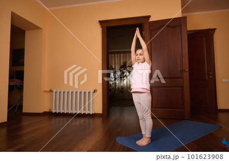Full length of a lovely little kid girl exercising on yoga mat, raising her arms up while stretching her body, practicing yoga, warming up while doing daily physical exercise at cozy homely atmosphere 101623908