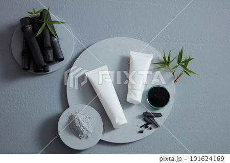 Top view of empty bottles for cosmetics, bamboo charcoal powder and finished products placed on a round tray. Bamboo charcoal and green leaves stand out against the gray background 101624169