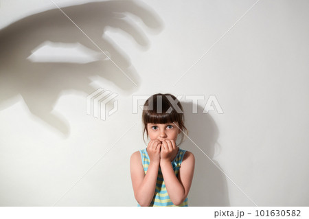 Scared small girl wearing casual sleeveless t shirt being afraid of shadows of hands on wall, looking at camera with frightened expression, isolated over gray background. Scared small girl wearing casual sleeveless t shirt being afraid of shadows of hands on wall, looking at camera with frightened expression, isolated over gray background. 101630582