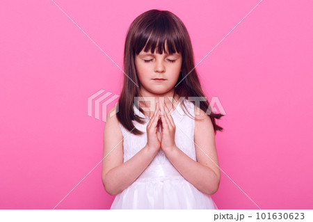 Calm beautiful little girl in white dress practicing yoga, keeping eyes closed, pressing palms together, dark haired cute kid praying, expressing hope, isolated over pink background. 101630623