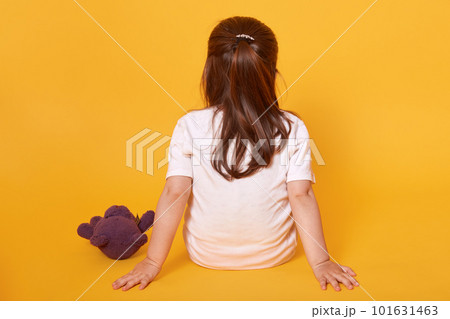 Rear view of little fair haired child sitting on the floor, putting hands aback, being punished, spending time alone, having brown teddy bear nearby, wearing white t shirt. Childhood concept. 101631463