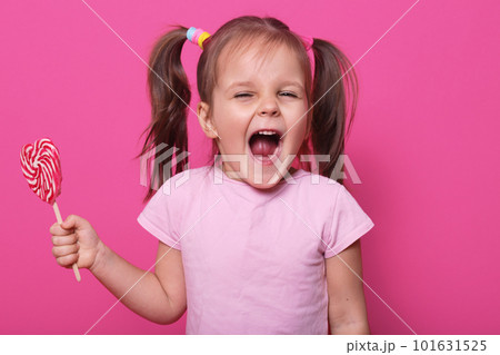 Studio shot of little attractive child opening mouth widely, looking directly at camera with excitement, holding bright lollipop in hand, wearing casually, isolated over pink studio background. Studio shot of little attractive child opening mouth widely, looking directly at camera with excitement, holding bright lollipop in hand, wearing casually, isolated over pink studio background. 101631525