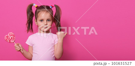 Close up portrait of little cute child, looking directly at camera, holding heart bright lollipop, pointing aside with her thumb, charming kid wearing casual rose t shirt, posing isolated on rose. Close up portrait of little cute child, looking directly at camera, holding heart bright lollipop, pointing aside with her thumb, charming kid wearing casual rose t shirt, posing isolated on rose. 101631527