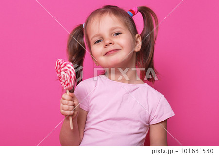 Close up portrait of cute child wearing rose casual t shirt, happy little girl holding big sugar lollipop, has gladness facial expression charming, posing isolated on pink background in studio. Close up portrait of cute child wearing rose casual t shirt, happy little girl holding big sugar lollipop, has gladness facial expression charming, posing isolated on pink background in studio. 101631530