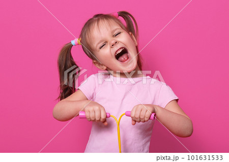 Adorable girl holds skipping rope in hands. Charmig female child with opened mouth and narrowed eyes with happy expression. LIttle cute model poses in photo studio isolated over pink bckground. 101631533