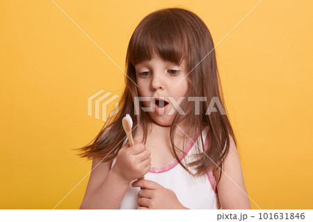 Studio shot of of cute little girl posing against yellow wall with toothbrush in hands, charming dark haired with opened mouth, being in shock, first teeth falling, strong toothache. Childhood concept 101631846