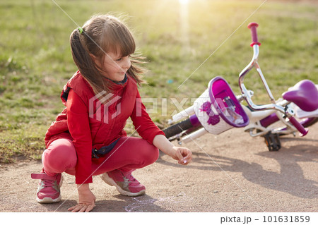 Charming little girl with ponytails dresses pink trousers and red shirt, drawing with chalk on asphalt, female kid being photographed outdoors near her bicycle.Childhood, children,spare time concept. 101631859