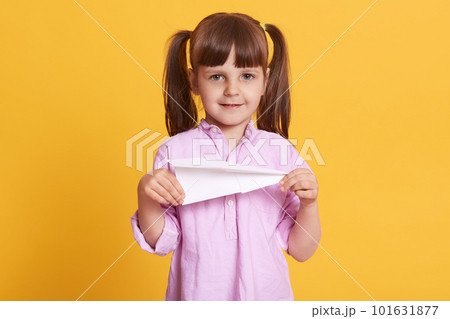 Image of delighted positive girl standing isolated over yellow background, holding paper plane in both hands, having fun, playing game, looking directly at camera, having peaceful facial expression. 101631877