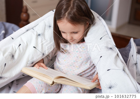 Portrait of charming child reading book at home, wrapped white blanket with dandelions, dark haired kid wearing pajama looking concentrated down on pages from her book. Children education concept. Portrait of charming child reading book at home, wrapped white blanket with dandelions, dark haired kid wearing pajama looking concentrated down on pages from her book. Children education concept. 101632775