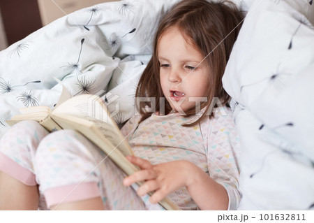 Close up portrait of little girl reading story or fairy tale for her teddy bears while laying in bed and wearing pajama, charming kid speling words aloud, looks concentrated, being in good mood. 101632811