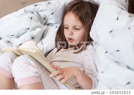 Profile of hard working curious smart girl learning to read, looking at text attentively, reading in voice, holding book, having fair hair, staying in bed, being on weekends at home. Study concept. Profile of hard working curious smart girl learning to read, looking at text attentively, reading in voice, holding book, having fair hair, staying in bed, being on weekends at home. Study concept. 101632813