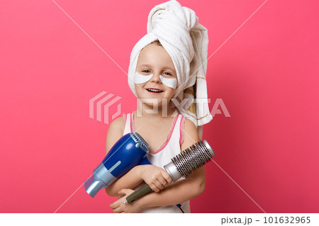 Horizontal shot of charming female kid posing isolated over rose studio background, little girl holding brush and hair dryer, kid making hairstyle, posing with towel on head and patches under eyes. 101632965