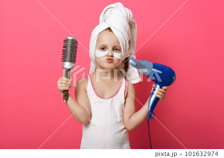 Picture of charming cute female kid isolated over rosy studio studio wall, girl being ready combing her hair with brush, making hairstyle, using hairdryer, adorable little lady keeping lips rounded. 101632974
