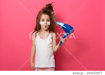 Portrait of pretty little girl with tangled hair comb in her hair, kid holding hair dryer and having astonished facial expression, has problem, model posing isolated on pink background. Kid's fashion. 101632977