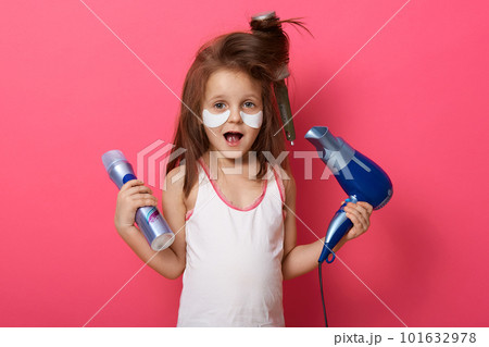 Portrait of charming cute female kid posing isolated over pink studio background, little girl combing her hair with brush, having problems, comb tangled while kid making hairstyle, using hairdryer. Portrait of charming cute female kid posing isolated over pink studio background, little girl combing her hair with brush, having problems, comb tangled while kid making hairstyle, using hairdryer. 101632978