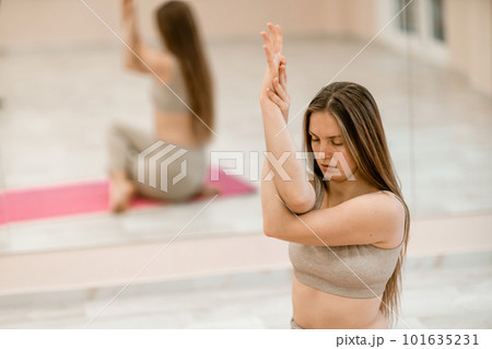 Young woman doing yoga in the gym. A girl with long hair and in a beige tracksuit stands in a cow pose on a pink carpet. A woman performs Gomukhasana. 101635231