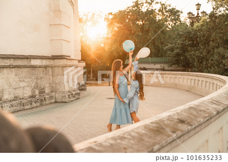 Daughter mother run holding hands. In blue dresses with flowing long hair, they hold balloons in their hands against the backdrop of a sunset and a white building. 101635233