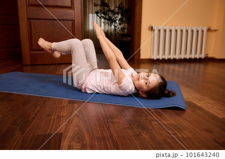 Happy relaxed Caucasian little child girl raising arms, doing abs exercises, stretching body, lying on a blue fitness mat and smiling looking at camera Happy relaxed Caucasian little child girl raising arms, doing abs exercises, stretching body, lying on a blue fitness mat and smiling looking at camera 101643240