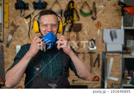 A carpenter in goggles and earmuffs removes a protective respirator. 101646895