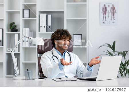Hindu doctor consulting patients remotely, man working inside modern clinic office, using laptop for online video call, talking and smiling friendly to patient. 101647947