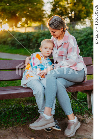 mother and son sit on a park bench in the rays of the setting sun. the concept of a family. Mother's Day. beautiful girl (mother) with a boy (son) in the park in the park are sitting on a bench at mother and son sit on a park bench in the rays of the setting sun. the concept of a family. Mother's Day. beautiful girl (mother) with a boy (son) in the park in the park are sitting on a bench at 101650926