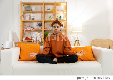 Yoga mindfulness meditation. Young healthy african girl practicing yoga at home. Woman sitting in lotus pose on couch meditating smiling relaxing indoor. Girl doing breathing practice. Yoga at home 101651185
