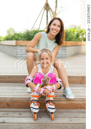 Little girl in roller skates and her mom sit on a wooden ladder and hug outdoors. Little girl in roller skates and her mom sit on a wooden ladder and hug outdoors. 101654965