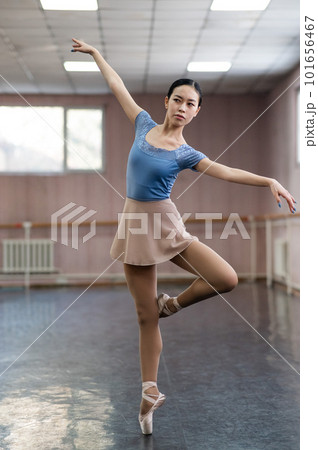 Graceful Asian ballerina in a blue bodysuit and a beige skirt is rehearsing in a dance class.  101656467