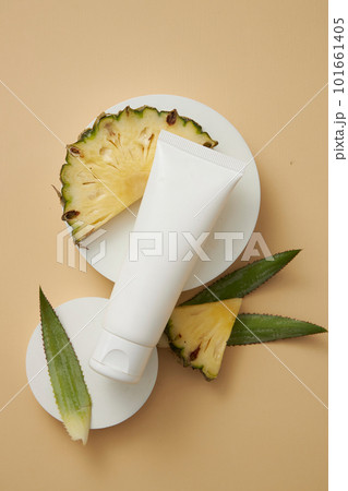 A white tube placed on round shaped podiums, displayed with pineapple leaves and slices. Empty label to promote beauty product extracted from pineapple (Ananas comosus) 101661405