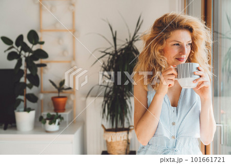 Waist up portrait woman with blonde curly hair holding a cup of tea standing by the window looking out, plants in the background Waist up portrait woman with blonde curly hair holding a cup of tea standing by the window looking out, plants in the background 101661721