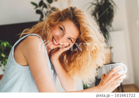 Cheerful curly woman looking at the camera with a big smile on her face wearing red lipstick and holding a cup of coffee in her hands 101661722