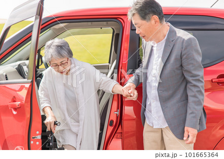 自動車の助手席から降りる杖をつく高齢者女性と介助する男性 101665364