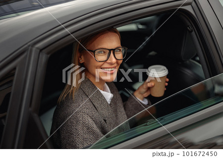 Smiling woman passenger in eyeglasses drinking takeaway coffee in taxi car on the way to work Smiling woman passenger in eyeglasses drinking takeaway coffee in taxi car on the way to work 101672450