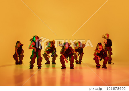 Dynamics. Group of children, little girls in sportive casual style clothes dancing hip-hop, contemp dance against yellow studio background in neon light Dynamics. Group of children, little girls in sportive casual style clothes dancing hip-hop, contemp dance against yellow studio background in neon light 101678492
