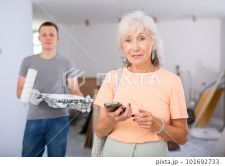 Satisfied grandmother inspects the room after the repair. In the background, a grandson with paint roller in his hands 101692733