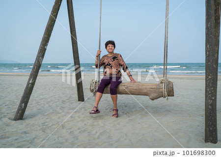 Senior Asian woman relaxing on a wood plank swing on the beach. 101698300