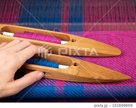 Hand takes a wooden boat shuttle with blue yarn to make a weaving project. Hand loom work 101699609