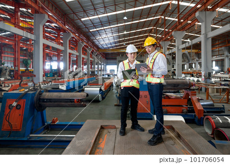 Young asian engineer manager trains new employees within the metal sheet factory. Everyone wear safety vest and hardhat. Various kind of industrial machine are in the working area. 101706054